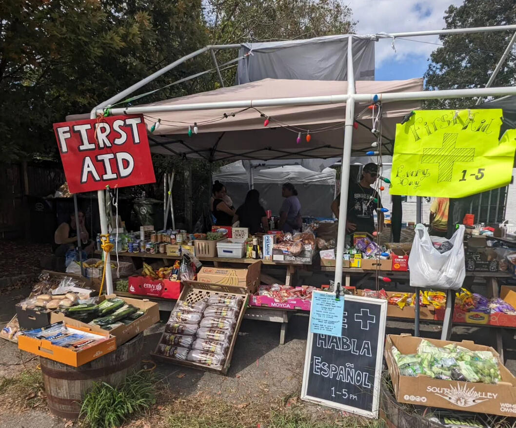 a pop-up tent, picnic tables and colorful signs outdoors. one sign reads "First Aid" and another reads "Se Habla Espanol"
